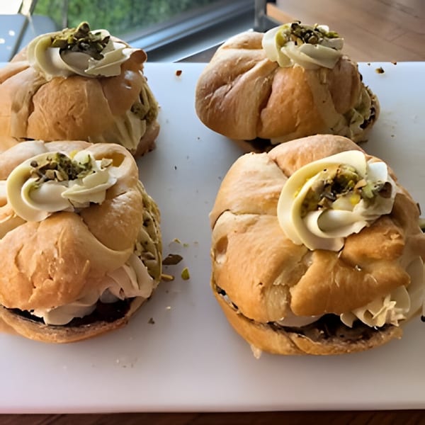 Four cream-filled pastries topped with pistachio crumbles and white chocolate curls on a white surface.