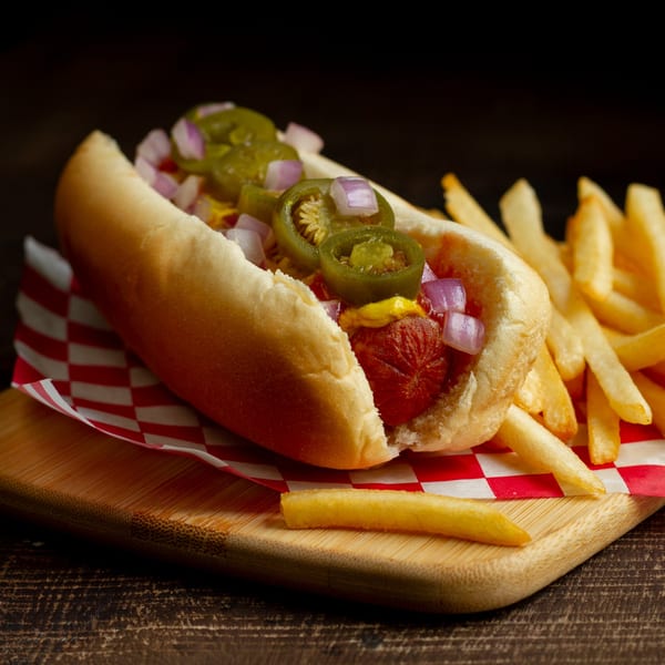 Hot dog with jalapeños, red onions, and mustard, served with fries on a checkered paper.