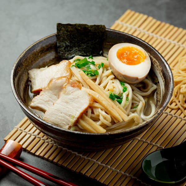 A bowl of ramen with pork, soft-boiled egg, bamboo shoots, and seaweed on a bamboo mat.